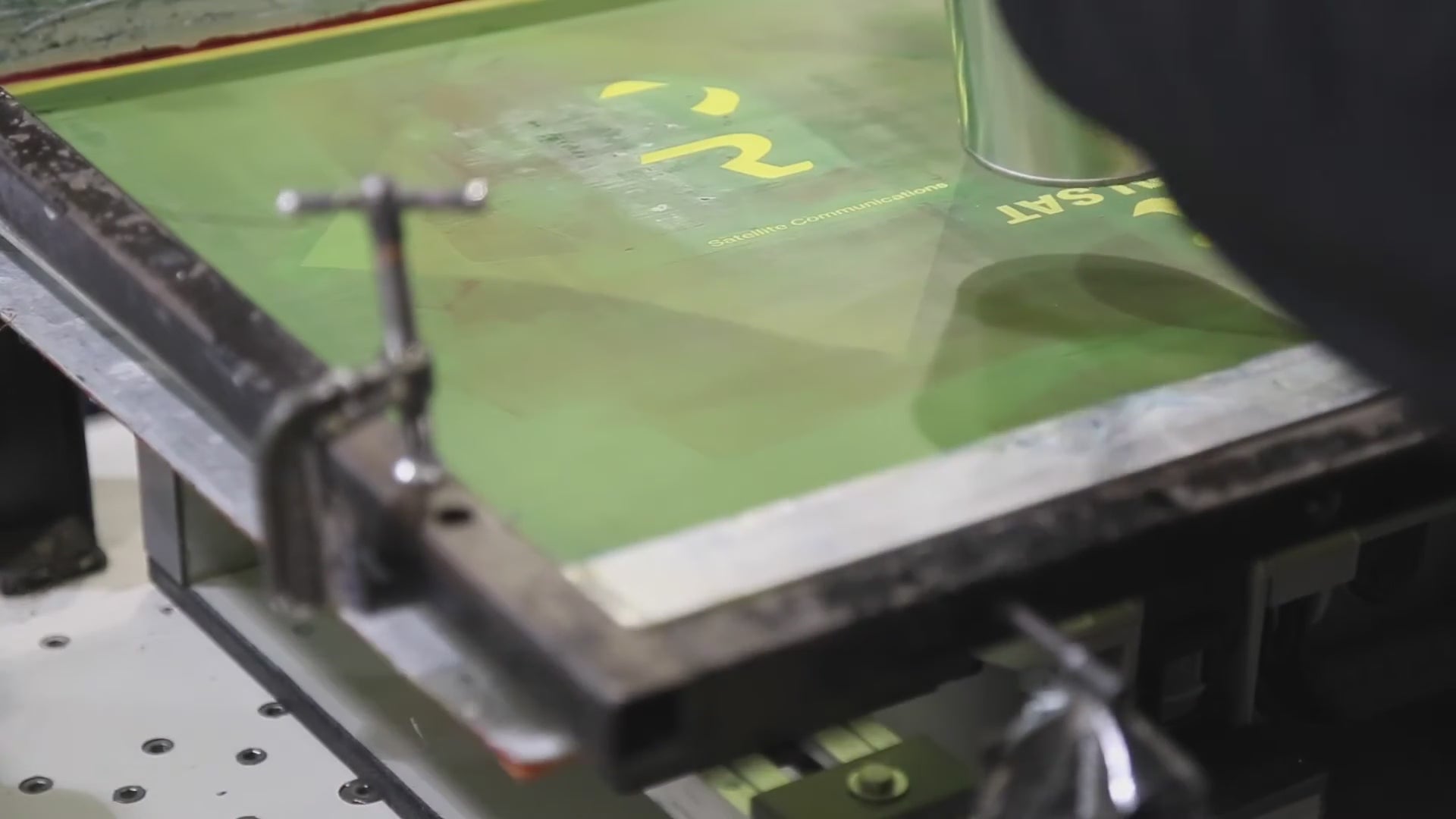 Load video: A close-up video demonstrates the multi-color screen printing process on a gray plastic product casing. A person applies successive layers of black, white, and bright green ink using a squeegee and stencil to precisely print the "REALSAT Satellite Communications" logo and a number "2" onto the component, showcasing detailed manual branding for durable goods.